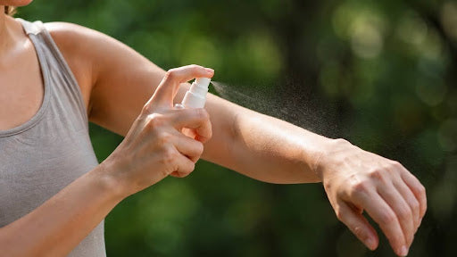 Woman spraying insect repellent on her forearm outdoors with green foliage in the background.